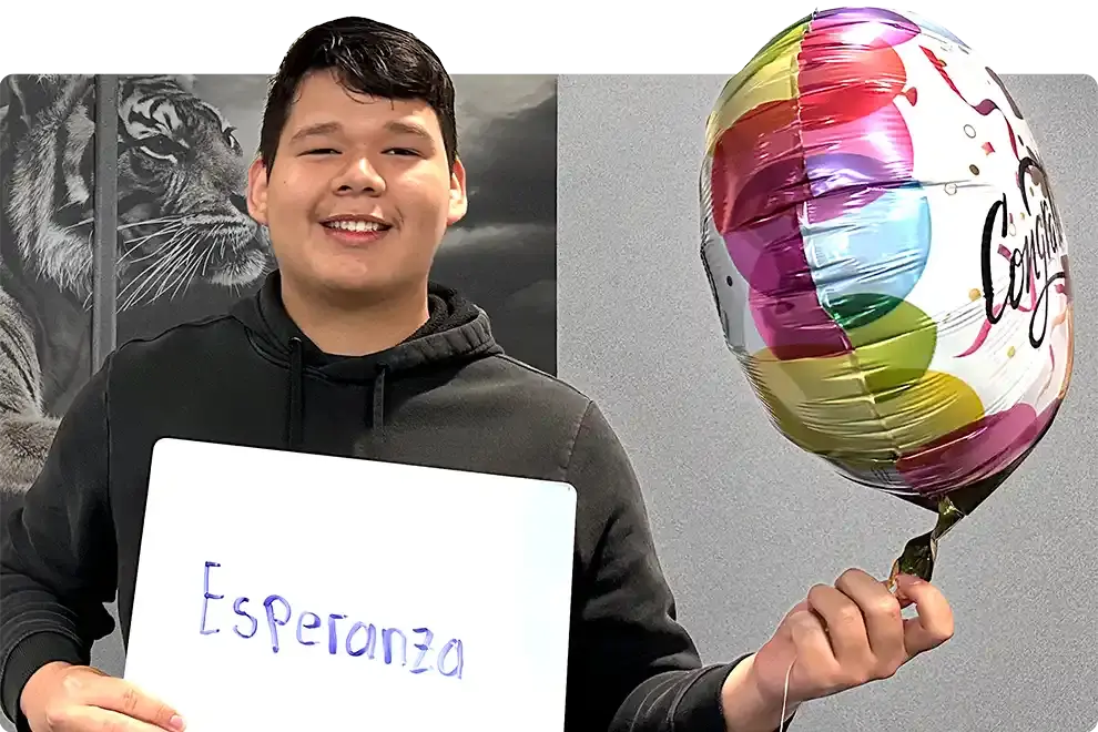 A smiling student holding a whiteboard on which he has written “Esperanza”, a Congratulations balloon in his other hand, in front of a painted tiger mural