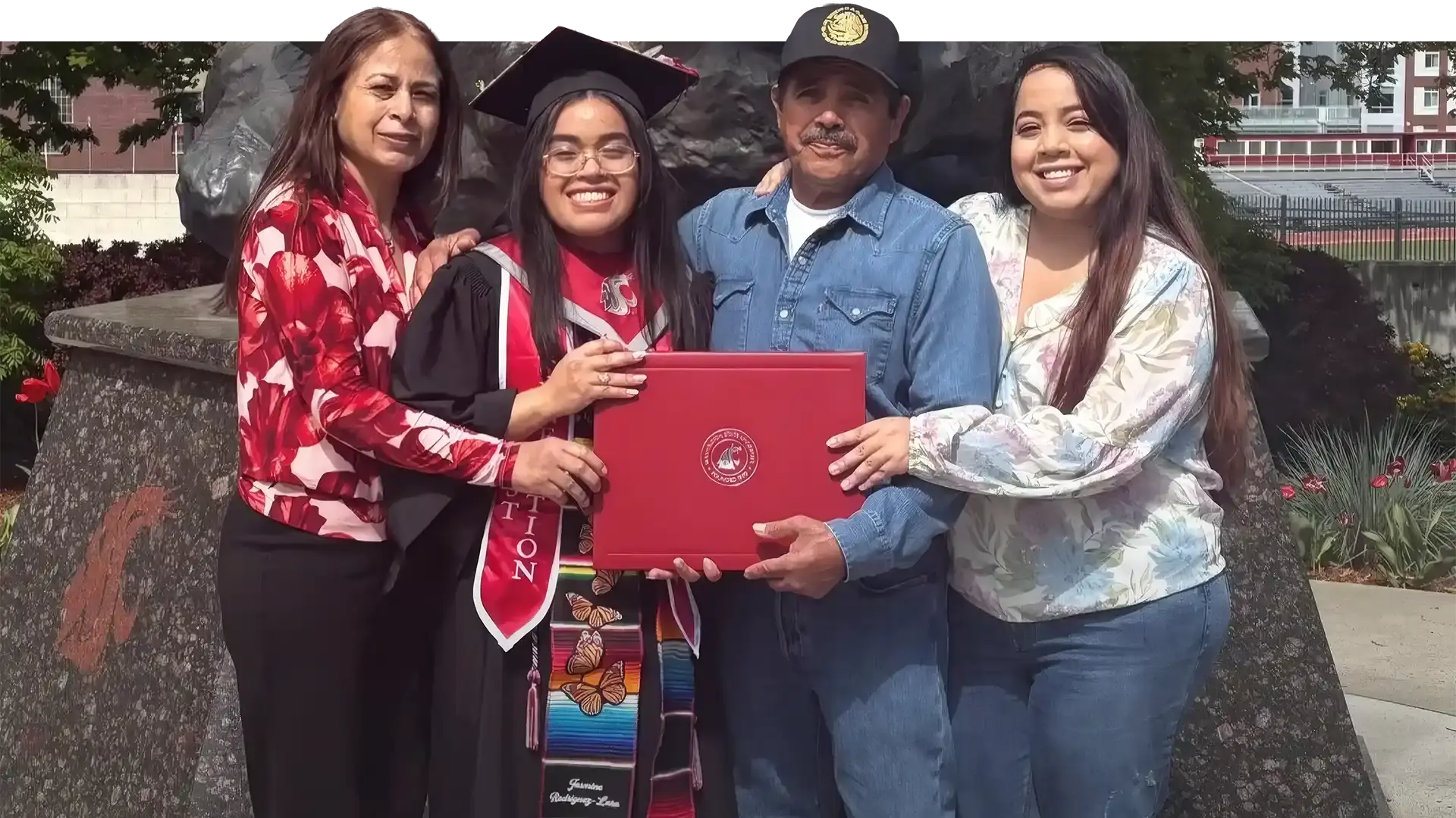 A WAEF graduate in cap and gown holding her diploma between her mother, her father and her sister, all four of them smiling at the camera on graduation day